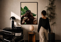 Black Man with Hand on Head print in a black frame on a sunlit plaster wall, a woman looking on beside a mushroom lamp