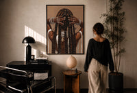 Black Woman Holding Her Braids print in a black frame on a sunlit plaster wall, a woman looking on beside a mushroom lamp