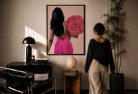 Black Woman Holding Pink Roses print in a black frame on a sunlit plaster wall, a woman looking on beside a mushroom lamp
