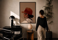 Black Woman in Red Baseball Cap print in a black frame on a sunlit plaster wall, a woman looking on beside a mushroom lamp