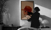 Black Woman in Red Baseball Cap print in a slim black frame being hung on a sunlit wall in a monochrome room