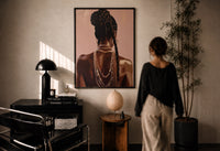 Black Woman with a Braided Bun print in a black frame on a sunlit plaster wall, a woman looking on beside a mushroom lamp