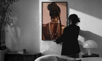 Black Woman with a Braided Bun print in a slim black frame being hung on a sunlit wall in a monochrome room