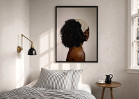Black Woman with a Curly Puff print in a black frame above a bed with striped linens, a brass wall lamp and a sunlit window
