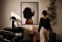 Black Woman with a Curly Puff print in a black frame on a sunlit plaster wall, a woman looking on beside a mushroom lamp