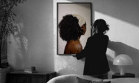 Black Woman with a Curly Puff print in a slim black frame being hung on a sunlit wall in a monochrome room