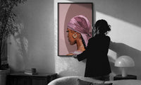 Black Woman with a Pink Headscarf print in a slim black frame being hung on a sunlit wall in a monochrome room