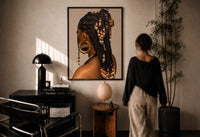 Black Woman with Beaded Braids print in a black frame on a sunlit plaster wall, a woman looking on beside a mushroom lamp