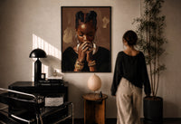 Black Woman with Clasped Hands print in a black frame on a sunlit plaster wall, a woman looking on beside a mushroom lamp