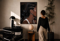 Black Woman with Cornrow Braids print in a black frame on a sunlit plaster wall, a woman looking on beside a mushroom lamp