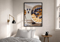 Blueberry Banana Oatmeal Bowl print in a black frame above a bed with striped linens, a brass wall lamp and a sunlit window