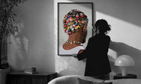 Bottle Cap Headpiece and Chunky Necklaces print in a slim black frame being hung on a sunlit wall in a monochrome room
