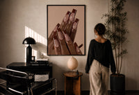 Brown French Tips print in a black frame on a sunlit plaster wall, a woman looking on beside a mushroom lamp