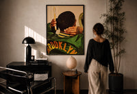 Brushing Waves in a Varsity Jacket print in a black frame on a sunlit plaster wall, a woman looking on beside a mushroom lamp