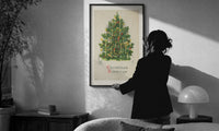 Christmas Tree Greetings print in a slim black frame being hung on a sunlit wall in a monochrome room