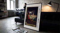 Man in red cloak holding lantern print in a slim black frame leaning on a dark sofa in a concrete print studio