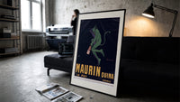 Green devil holding aperitif bottle print in a slim black frame leaning on a dark sofa in a concrete print studio