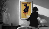 Cappiello Parapluie-Revel figures print in a slim black frame being hung on a sunlit wall in a monochrome room