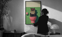 Cat on the Toilet Reading the Paper print in a slim black frame being hung on a sunlit wall in a monochrome room