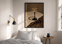 Champagne Coupe with Leopard print in a black frame above a bed with striped linens, a brass wall lamp and a sunlit window