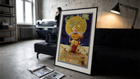 Pumpkin-headed figure with harvest moon print in a slim black frame leaning on a dark sofa in a concrete print studio