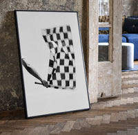 Hand waving checkered racing flag print in a black frame leaning beside a vintage door on parquet