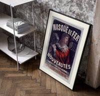 Man in red cloak holding iron mask poster in a black frame leaning beside a shelf in a raw-plaster loft