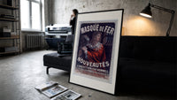 Man in red cloak holding iron mask print in a slim black frame leaning on a dark sofa in a concrete print studio