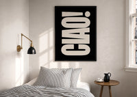 Ciao print in a black frame above a bed with striped linens, a brass wall lamp and a sunlit window