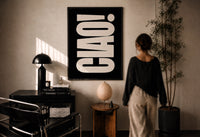 Ciao print in a black frame on a sunlit plaster wall, a woman looking on beside a mushroom lamp