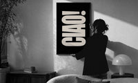 Ciao print in a slim black frame being hung on a sunlit wall in a monochrome room