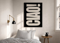 Ciao White on Black print in a black frame above a bed with striped linens, a brass wall lamp and a sunlit window