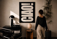 Ciao White on Black print in a black frame on a sunlit plaster wall, a woman looking on beside a mushroom lamp