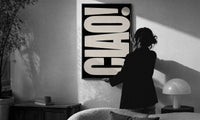 Ciao White on Black print in a slim black frame being hung on a sunlit wall in a monochrome room