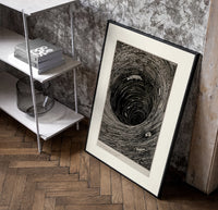 Boat caught in whirlpool vortex poster in a black frame leaning beside a shelf in a raw-plaster loft