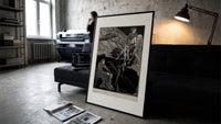 Man carrying pale woman, seated figure print in a slim black frame leaning on a dark sofa in a concrete print studio