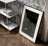 Two figures in boat amid rocks poster in a black frame leaning beside a shelf in a raw-plaster loft