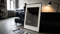 Two figures in boat amid rocks print in a slim black frame leaning on a dark sofa in a concrete print studio
