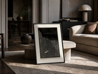 Two figures in boat amid rocks print in a black frame leaning on a travertine table in a sunlit lounge