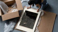Two figures in boat amid rocks framed print being unpacked from protective bubble wrap and a box