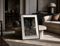 Robed figure atop dark mound print in a black frame leaning on a travertine table in a sunlit lounge