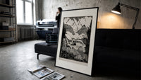 Mountain valley landscape with figure print in a slim black frame leaning on a dark sofa in a concrete print studio