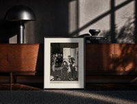 Black-framed seated man examined by standing figure print on a moody console with designer coffee-table books