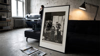 seated man examined by standing figure print in a slim black frame leaning on a dark sofa in a concrete print studio