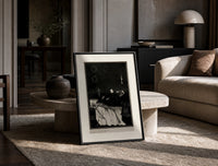 Decaying corpse on bed with figures print in a black frame leaning on a travertine table in a sunlit lounge
