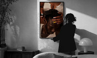 Combing Hair with Painted Nails print in a slim black frame being hung on a sunlit wall in a monochrome room