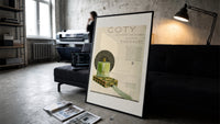Green perfume bottle on box print in a slim black frame leaning on a dark sofa in a concrete print studio
