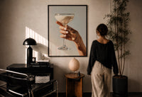 Cream Coupe Cocktail print in a black frame on a sunlit plaster wall, a woman looking on beside a mushroom lamp