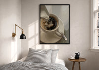 Cream Poured into Black Coffee print in a black frame above a bed with striped linens, a brass wall lamp and a sunlit window