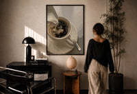 Cream Poured into Black Coffee print in a black frame on a sunlit plaster wall, a woman looking on beside a mushroom lamp